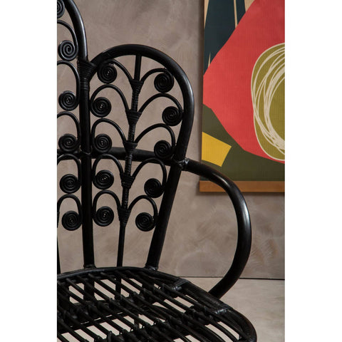 A black rattan chair with a scalloped back design featuring intricate leaf patterns, positioned against a textured wall and a colorful abstract artwork in the background. The chair's armrests are visible, showcasing its elegant curves