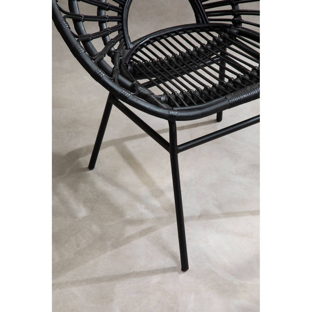 A black rattan flared back chair with a circular seat and open weave design, supported by slender black metal legs, placed on a light-colored floor