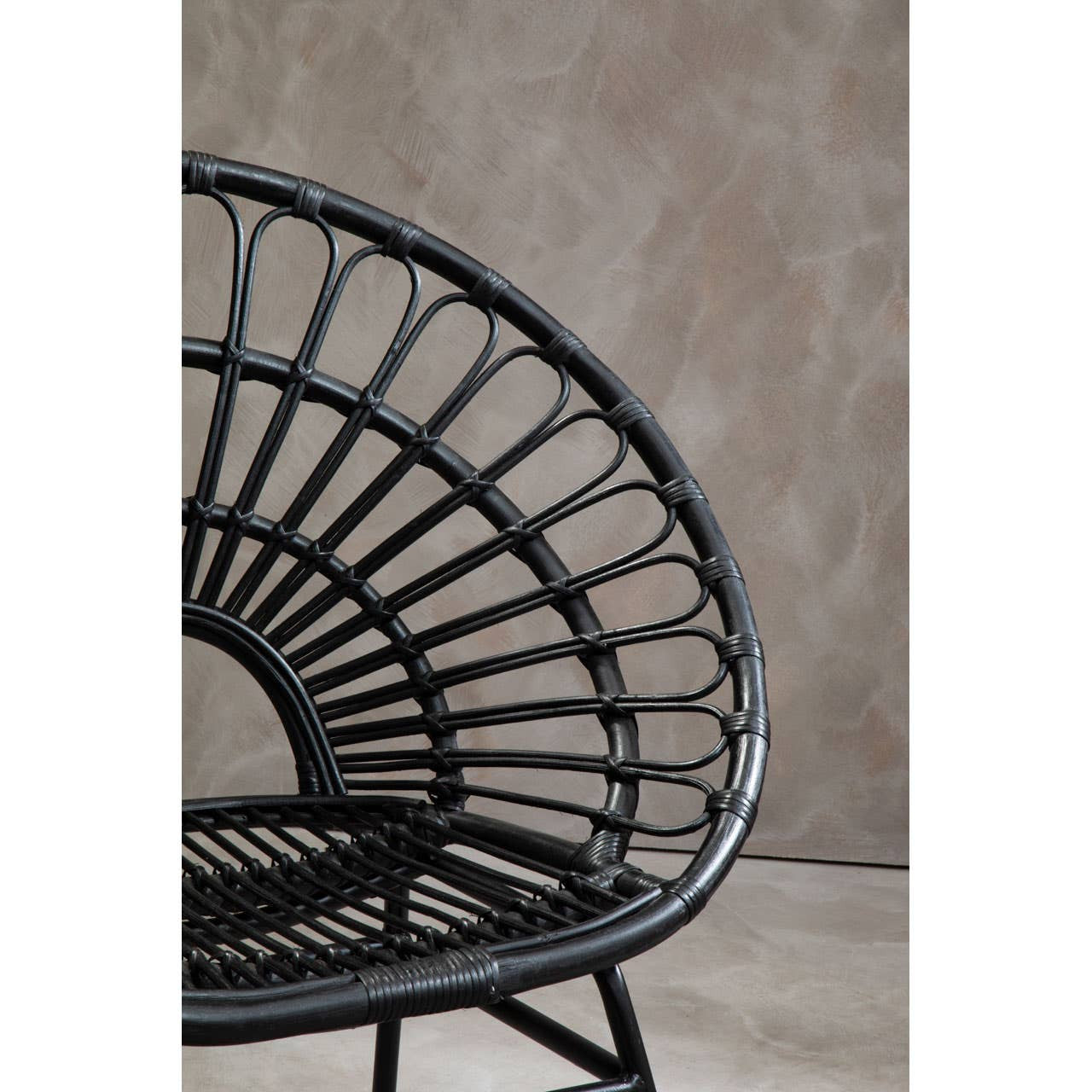 A flared back chair made of black natural rattan, featuring a circular woven design with a sturdy metal frame. The chair's backrest has a distinctive fan shape, and the seat is open and airy, set against a neutral textured background