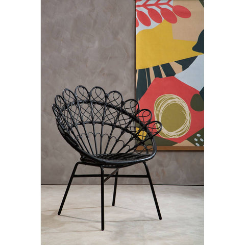 A black natural rattan peacock chair with a distinctive fan-shaped backrest, featuring curved lines and a sturdy metal frame. The chair is positioned on a light-colored floor against a textured gray wall, with a colorful abstract artwork in the background