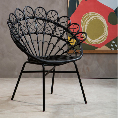 A stylish black rattan peacock chair with a distinctive fan-shaped backrest, supported by slender metal legs, set against a textured gray wall and colorful abstract artwork in the background