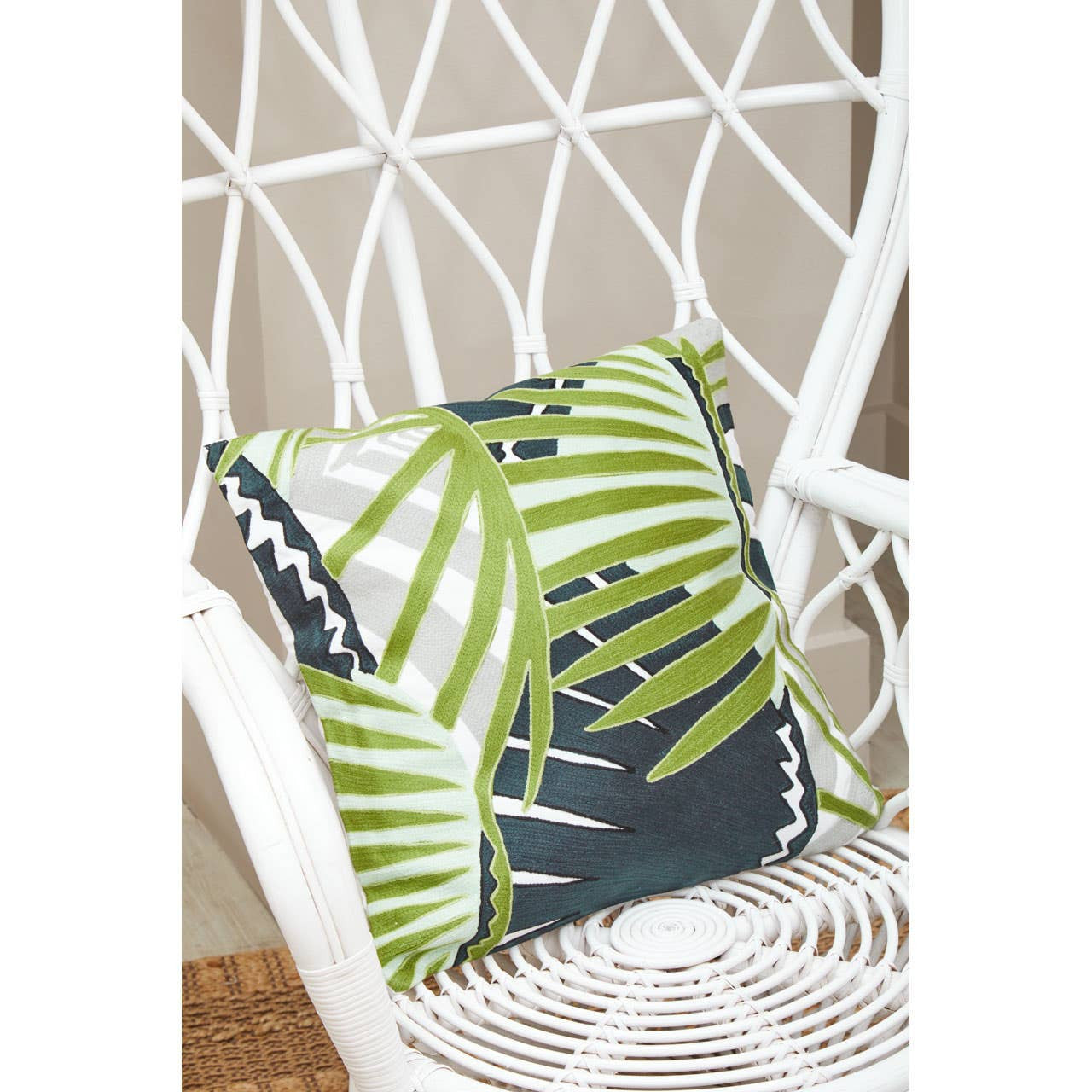 Curved chair made of natural rattan featuring a grey and white design, with a decorative green and navy blue leaf-patterned cushion placed on the seat. The background includes a light-colored wall and a woven mat on the floor