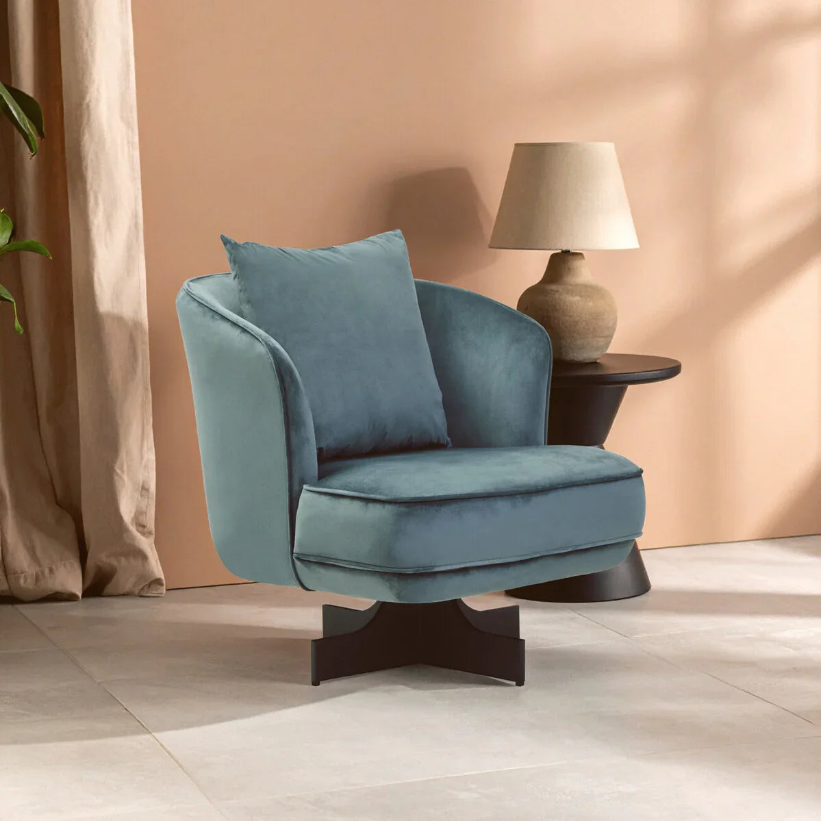 Blue velvet swivel armchair with cushion beside round black side table and beige lamp