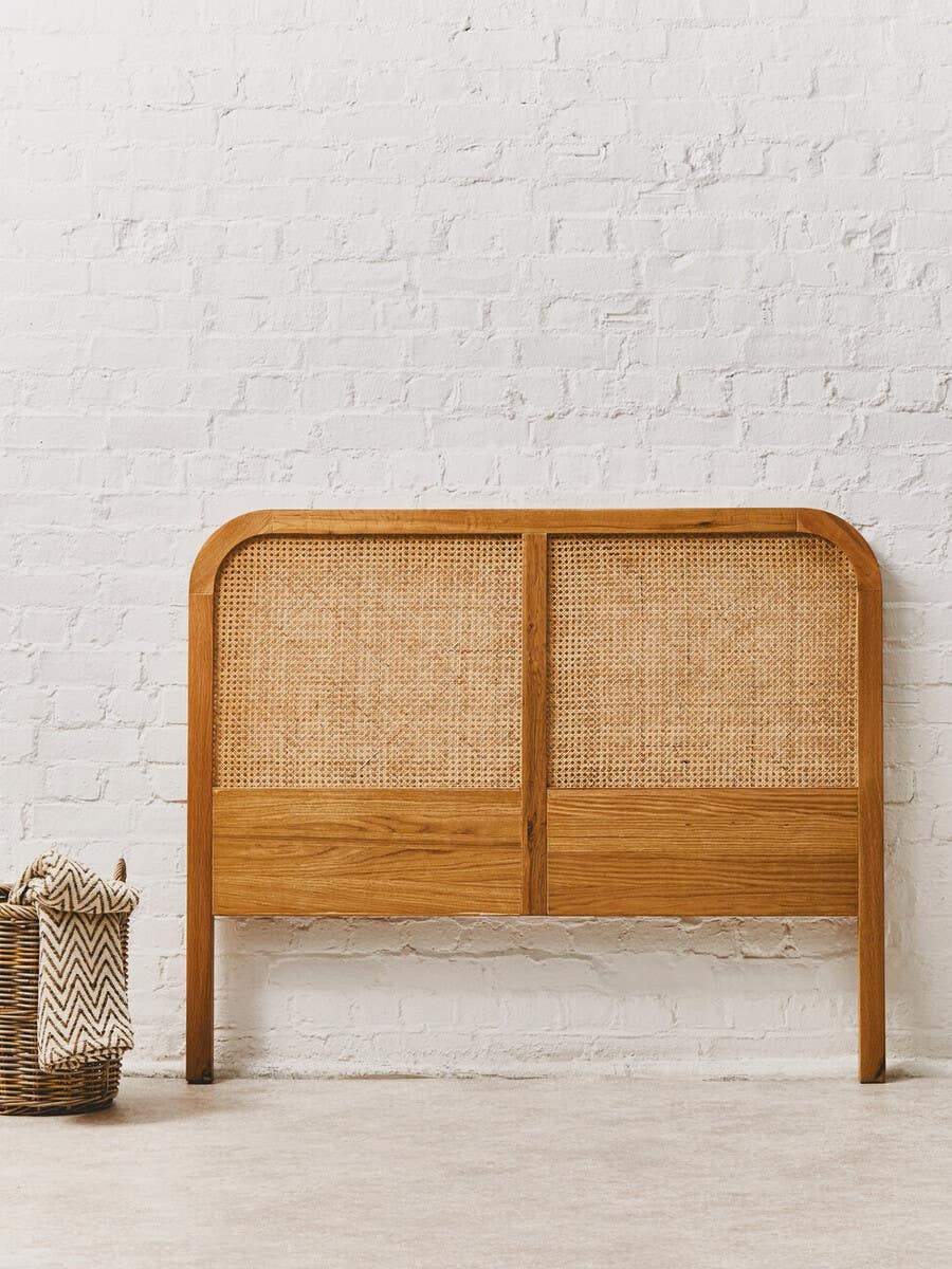 Lyon Rattan and Oak Wood Double Bed Headboard featuring a curved top and a combination of natural rattan and oak wood, displayed against a textured white wall with a light-colored floor. A small wooden crate is positioned to the left of the headboard