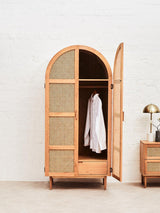 Lyon Rattan and Oak Wood 2 Door Wardrobe featuring a curved top, natural oak frame, and rattan paneling. One door is open, revealing a hanging white garment inside. The wardrobe stands on a light-colored floor with a bedside table and lamp partially visible in the background