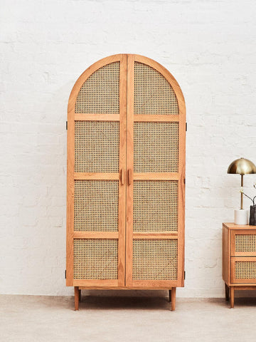 Lyon Rattan and Oak Wood 2 Door Wardrobe featuring a curved top, natural rattan front with woven patterns, and oak wood frame. The wardrobe has two doors and stands on short legs, with a lamp and a small side table visible beside it. The background is a neutral wall, enhancing the wardrobe's design
