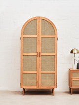 Lyon Rattan and Oak Wood 2 Door Wardrobe featuring a curved top, natural rattan front with woven patterns, and oak wood frame. The wardrobe has two doors and stands on short legs, with a lamp and a small side table visible beside it. The background is a neutral wall, enhancing the wardrobe's design