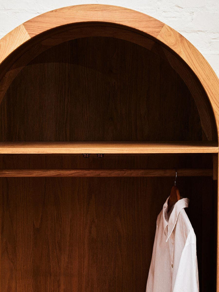 The interior of a wardrobe featuring a smooth wooden shelf and a hanging space with a white garment. The wardrobe has a rounded top design and is made from oak wood and rattan. The background is a light-colored wall, enhancing the natural tones of the wood