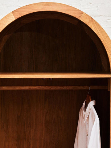 The interior of a wardrobe featuring a smooth wooden shelf and a hanging space with a white garment. The wardrobe has a rounded top design and is made from oak wood and rattan. The background is a light-colored wall, enhancing the natural tones of the wood