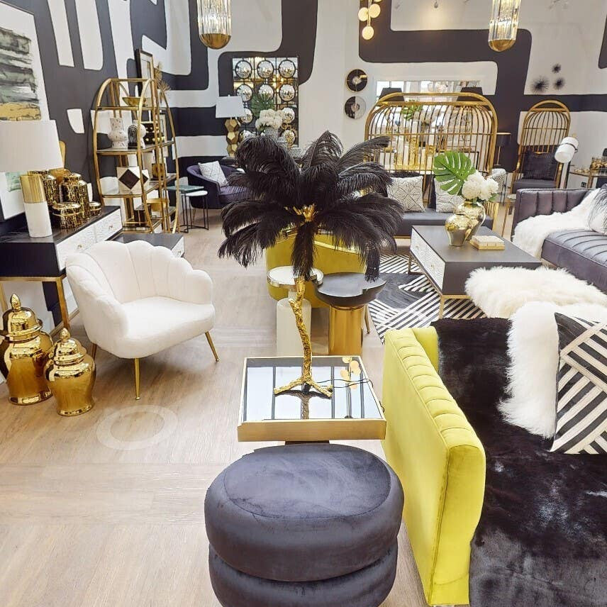 A black velvet oval pouffe sits on a wooden floor, surrounded by various pieces of modern furniture, including a white chair and a yellow accent table. The pouffe features a smooth texture and a rounded shape, complementing the contemporary decor of the space