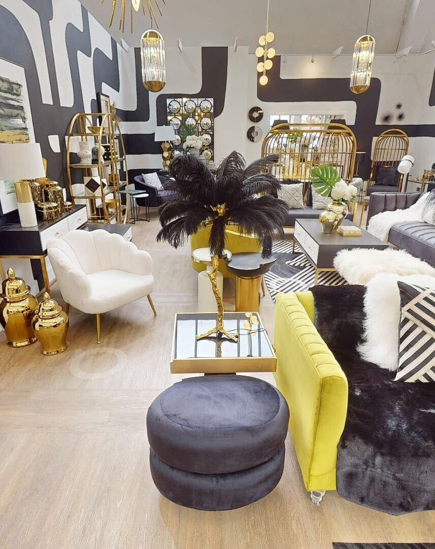 A black velvet oval pouffe sits on a wooden floor, surrounded by various pieces of modern furniture, including a white chair and a yellow accent table. The pouffe features a smooth texture and a rounded shape, complementing the contemporary decor of the space
