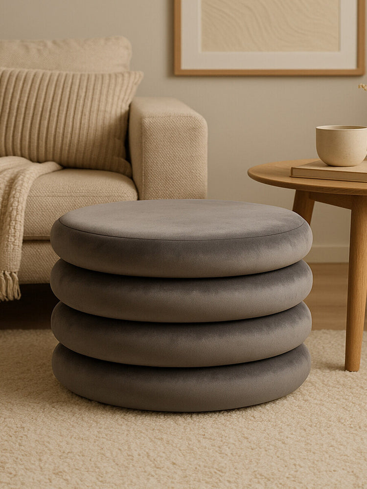 Round pouffe in grey velvet with a smooth, soft texture, featuring a stacked design of four circular layers. The pouffe is placed on a light-colored carpet, next to a wooden side table and a cozy armchair with a knitted throw draped over it. A neutral-toned wall art piece is visible in the background