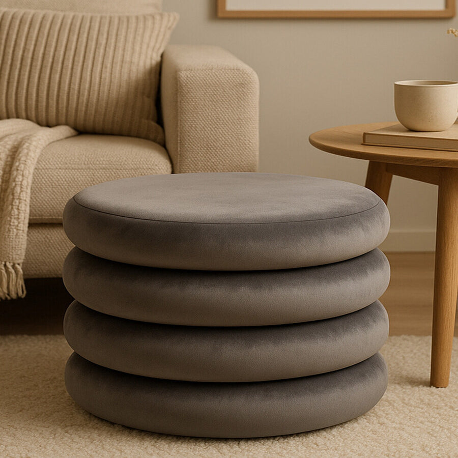 Round pouffe in grey velvet with a smooth, soft texture, featuring a stacked design of four circular layers. The pouffe is placed on a light-colored carpet, next to a wooden side table and a cozy armchair with a knitted throw draped over it. A neutral-toned wall art piece is visible in the background
