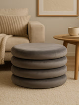 Round pouffe in grey velvet with a smooth, soft texture, featuring a stacked design of four circular layers. The pouffe is placed on a light-colored carpet, next to a wooden side table and a cozy armchair with a knitted throw draped over it. A neutral-toned wall art piece is visible in the background