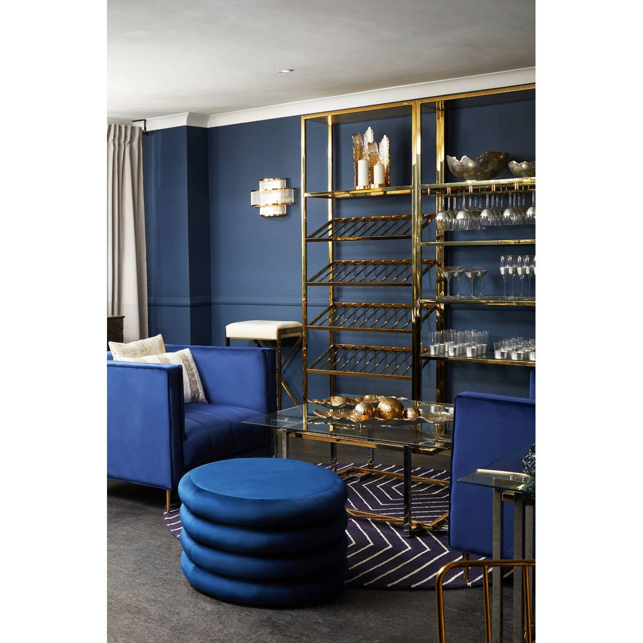 A round pouffe in midnight navy velvet is placed on a dark floor, surrounded by modern furniture in a stylish living space. The backdrop features a deep blue wall and a gold shelving unit displaying decorative items. Two blue upholstered chairs are visible, creating a cohesive color scheme