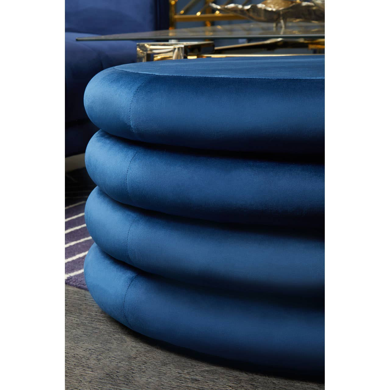 Round pouffe in midnight navy velvet, featuring a smooth, plush surface with a stacked design. The pouffe is set against a textured rug, showcasing its rich color and soft texture