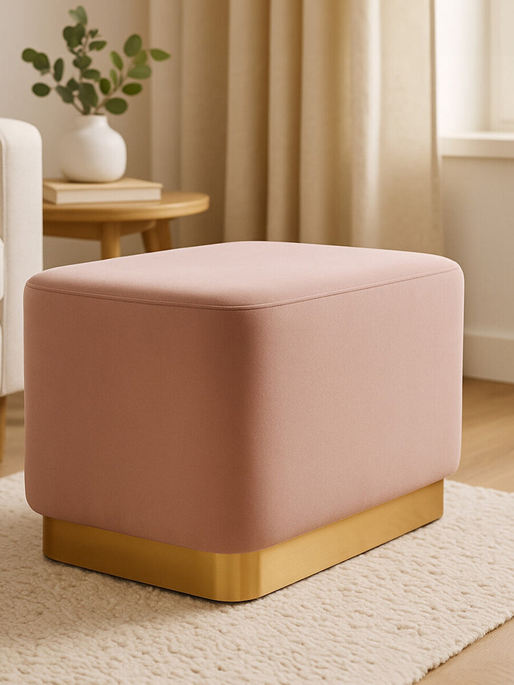 Dusty pink velvet rectangular pouffe with a matte gold base, positioned on a light-colored rug in a softly lit room with natural light filtering through a window