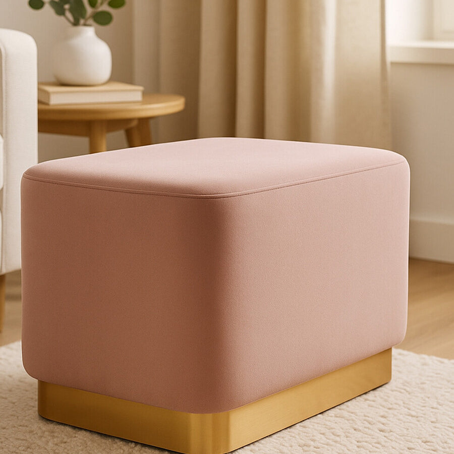 Dusty pink velvet rectangular pouffe with a matte gold base, positioned on a light-colored rug in a softly lit room with natural light filtering through a window