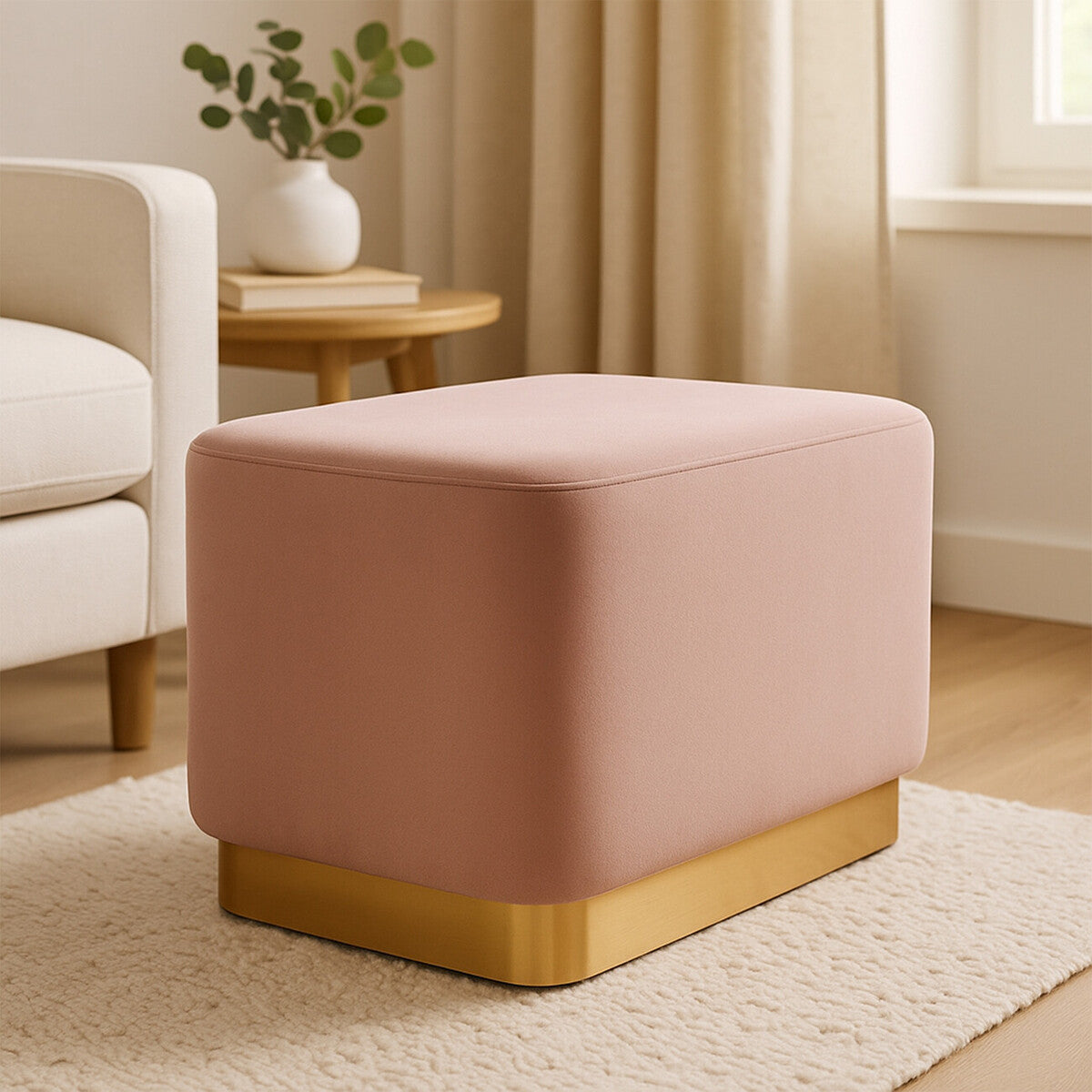 Dusty pink velvet rectangular pouffe with a matte gold base, placed on a light-colored rug in a cozy living room setting. Soft natural light filters through a nearby window, illuminating the pouffe and surrounding furniture