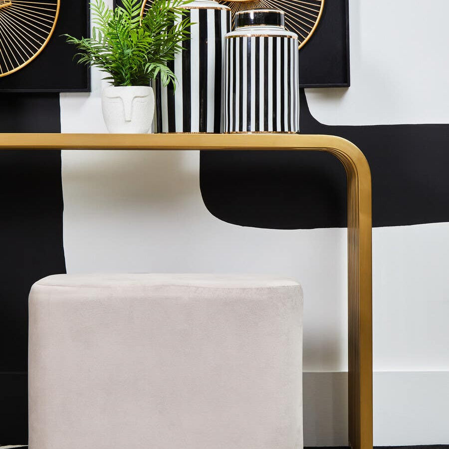A rectangular pouffe upholstered in soft mink velvet, featuring a matte gold base. The pouffe is positioned next to a modern table with a curved design, accompanied by decorative vases and a small green plant in the background. The overall color scheme includes neutral tones and metallic accents, creating a stylish and contemporary look