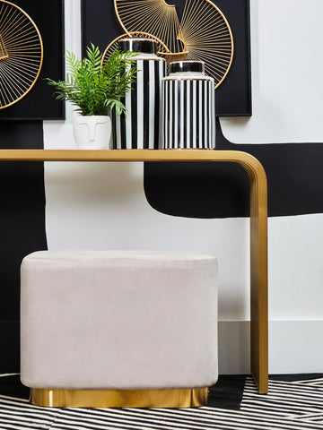 A rectangular pouffe upholstered in soft mink velvet, featuring a matte gold base. The pouffe is positioned next to a modern table with a curved design, accompanied by decorative vases and a small green plant in the background. The overall color scheme includes neutral tones and metallic accents, creating a stylish and contemporary look