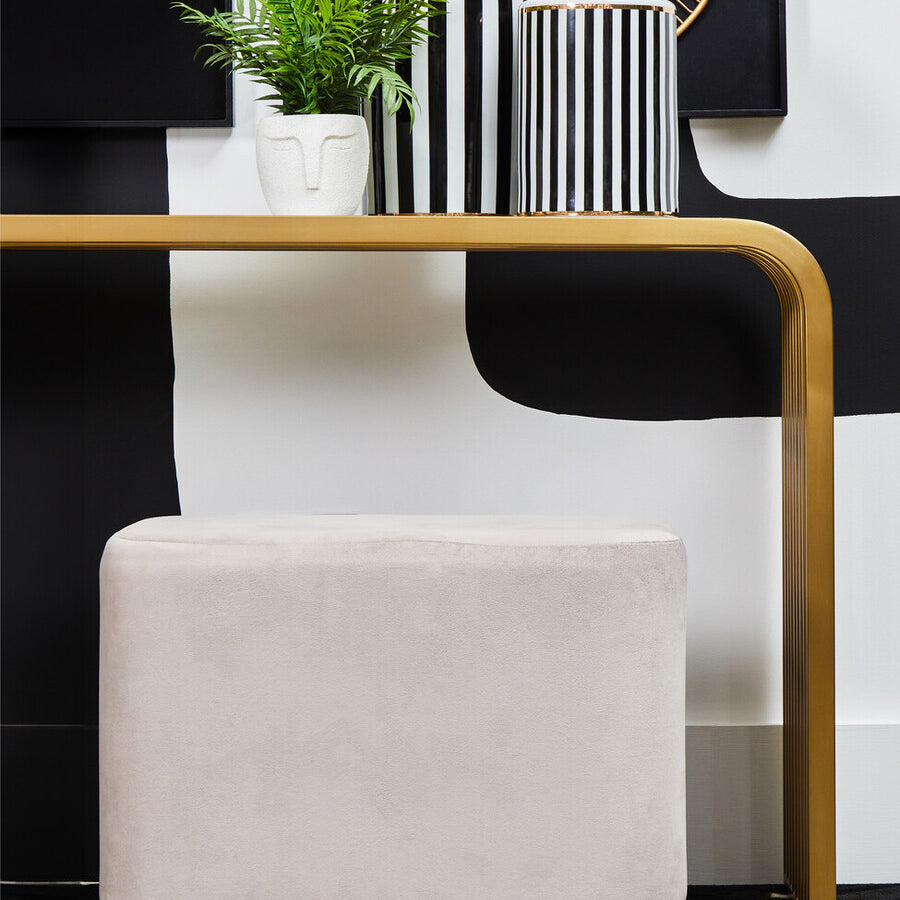 A rectangular pouffe upholstered in soft mink velvet with a matte gold base, positioned next to a modern console table. The pouffe features clean lines and a minimalist design, complementing the contemporary decor, which includes decorative vases and a small plant