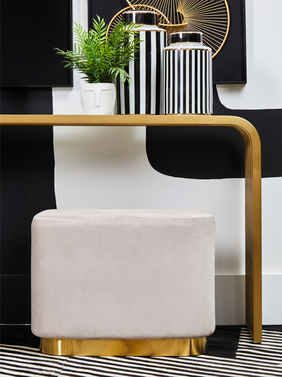 A rectangular pouffe upholstered in soft mink velvet with a matte gold base, positioned next to a modern console table. The pouffe features clean lines and a minimalist design, complementing the contemporary decor, which includes decorative vases and a small plant