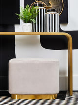 A rectangular pouffe upholstered in soft mink velvet with a matte gold base, positioned next to a modern console table. The pouffe features clean lines and a minimalist design, complementing the contemporary decor, which includes decorative vases and a small plant