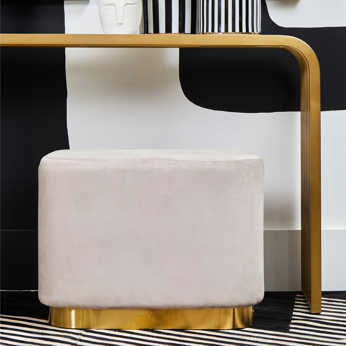 Rectangular pouffe upholstered in soft mink velvet with a matte gold base, positioned near a modern black and white furniture piece. The pouffe features rounded edges and a plush surface, complementing the sleek design of the surrounding decor