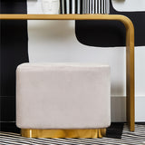 Rectangular pouffe upholstered in soft mink velvet with a matte gold base, positioned near a modern black and white furniture piece. The pouffe features rounded edges and a plush surface, complementing the sleek design of the surrounding decor
