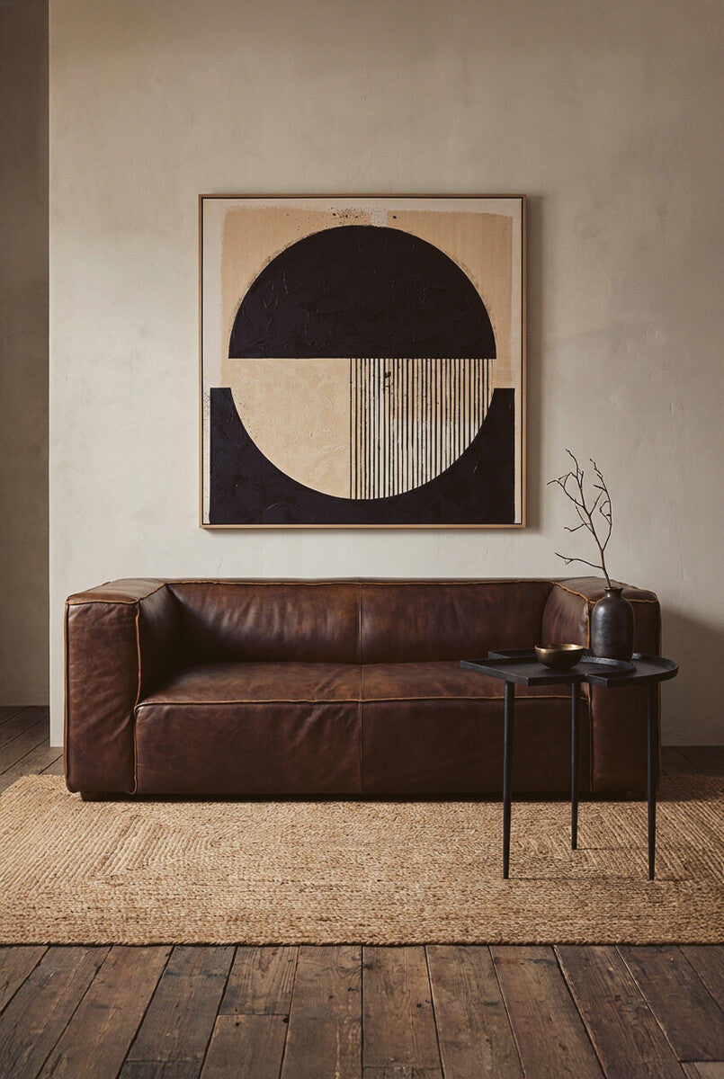 Antique brown leather two-seater sofa with a minimalist design, placed against a textured wall. A large abstract artwork featuring geometric shapes hangs above the sofa, and a small side table with a glass and a decorative item is positioned next to it. The floor is covered with a woven rug, adding warmth to the setting