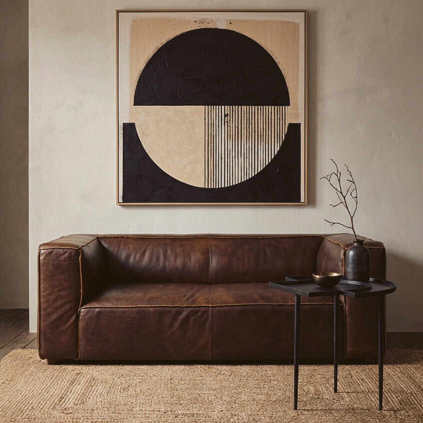 Antique brown leather two-seater sofa with a minimalist design, featuring clean lines and a plush seat. The sofa is positioned against a neutral wall, complemented by a large abstract artwork above it. A small black side table with a decorative vase sits next to the sofa, and the floor is covered with light wooden planks