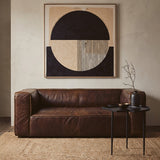 Antique brown leather two-seater sofa with a minimalist design, featuring clean lines and a plush seat. The sofa is positioned against a neutral wall, complemented by a large abstract artwork above it. A small black side table with a decorative vase sits next to the sofa, and the floor is covered with light wooden planks