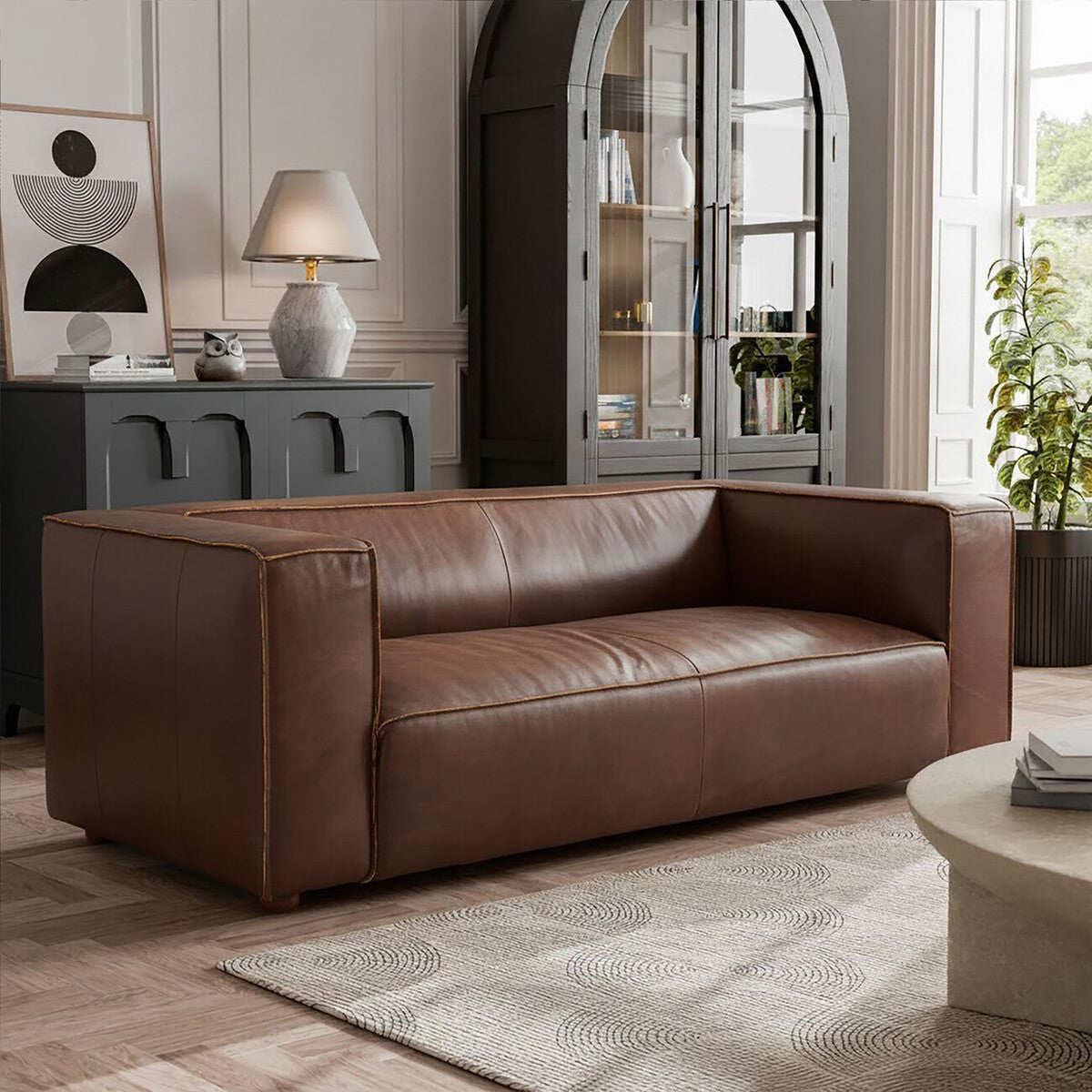 Antique brown leather two-seater sofa with a modern design, featuring clean lines and a low backrest. The sofa is placed on a light-colored rug in a well-lit room with a large window, a decorative lamp, and a stylish cabinet in the background