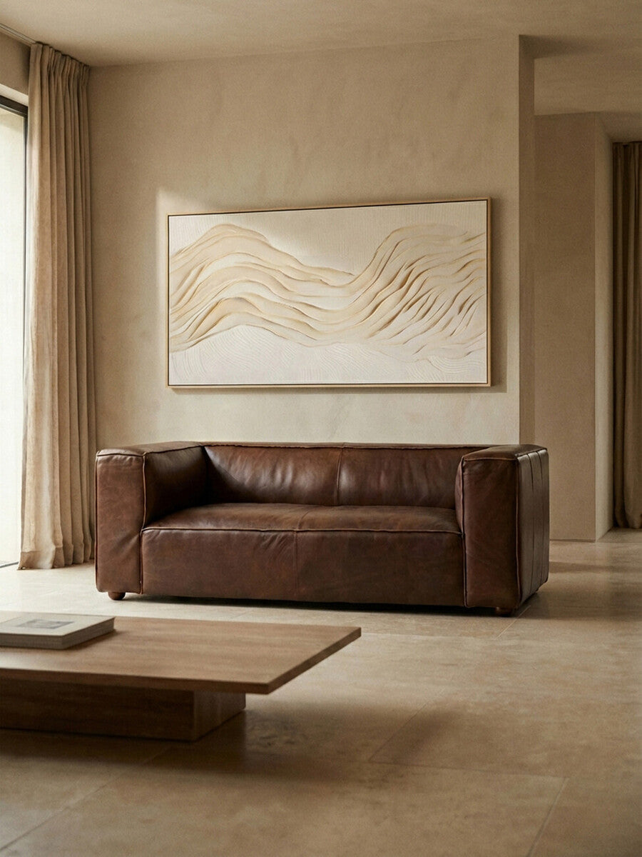 Antique brown leather two-seater sofa with a minimalist design, positioned against a neutral-colored wall. A large piece of abstract art hangs above the sofa, and a wooden coffee table is placed in front, complementing the warm tones of the room. Natural light filters in through a window, enhancing the cozy atmosphere