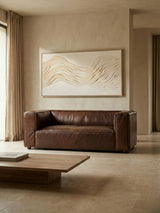 Antique brown leather two-seater sofa with a minimalist design, positioned against a neutral-colored wall. A large piece of abstract art hangs above the sofa, and a wooden coffee table is placed in front, complementing the warm tones of the room. Natural light filters in through a window, enhancing the cozy atmosphere