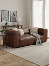 Antique brown leather two-seater sofa with a modern design, featuring plush cushions and a sleek silhouette. The sofa is positioned on a light-colored area rug, with a wooden side table beside it, adorned with a lamp and decorative items. Two framed artworks are displayed on the wall in the background, and natural light filters through sheer curtains