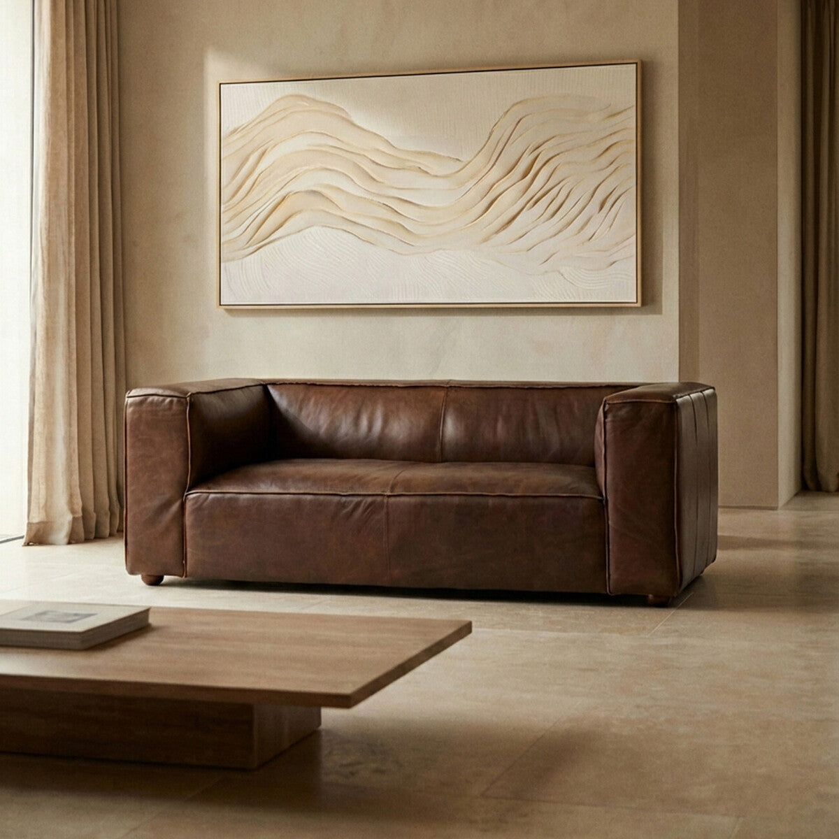 Antique brown leather two-seater sofa with a modern design, featuring thick armrests and a plush seat, positioned in a softly lit room with neutral-toned walls and a large abstract artwork above it. A wooden coffee table is placed in front of the sofa on a light-colored floor