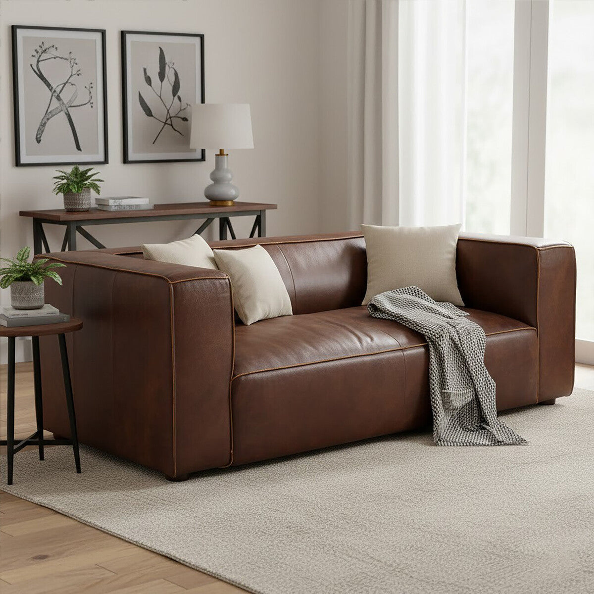 Antique brown leather two-seater sofa with a modern design, featuring thick armrests and a plush seat. A light-colored throw blanket is draped over one side, and two cream-colored cushions are placed on the seat. In the background, there is a wooden table with a decorative lamp and two framed artworks on the wall. The setting includes a light-colored rug and large windows allowing natural light to