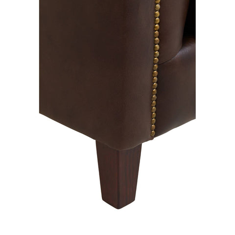 Close-up view of the leg and side of a dark brown leather Chesterfield sofa, showcasing the rich texture of the leather and decorative gold nailhead trim along the edge. The leg is wooden and tapered, providing a modern touch to the classic design