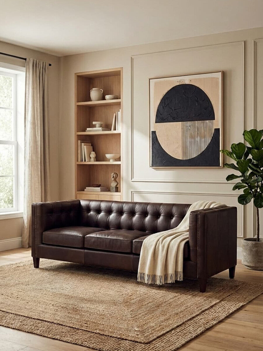 A dark brown leather Chesterfield sofa with a modern design, featuring tufted upholstery and a cozy throw blanket draped over one arm. The sofa is positioned in a bright room with large windows, a wooden bookshelf displaying decorative items, and a piece of abstract wall art. A textured area rug is placed underneath the sofa, adding warmth to the space