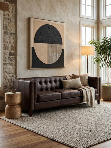A dark brown leather Chesterfield sofa with a modern design, featuring tufted upholstery and three seating sections. The sofa is positioned in a well-lit room with large windows, a textured area rug underneath, and a decorative wall art piece featuring geometric shapes in neutral tones. A floor lamp and a potted plant are also visible in the background, adding to the cozy atmosphere