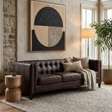 A dark brown leather Chesterfield sofa with a tufted backrest and rolled arms, positioned in a cozy living room setting. The sofa is accompanied by decorative pillows and a soft throw blanket. A large abstract wall art piece hangs above the sofa, and a floor lamp illuminates the space. Two round wooden side tables are placed beside the sofa, and a textured area rug covers the floor. Natural light