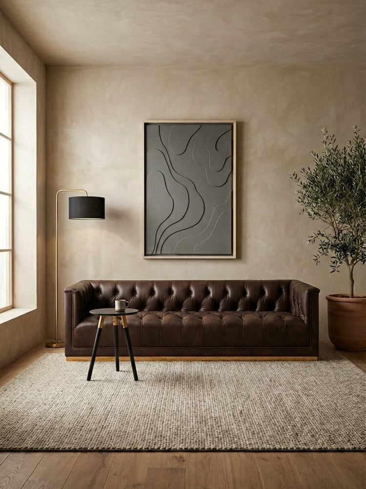 A dark brown leather Chesterfield sofa with a tufted backrest and armrests, positioned against a neutral-toned wall. A large abstract artwork hangs above the sofa, and a floor lamp stands beside it. A potted plant is placed nearby, and a textured area rug covers the floor. Natural light streams in through a large window, illuminating the scene