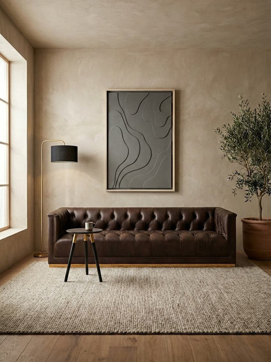 A dark brown leather Chesterfield sofa with a tufted backrest and armrests, positioned against a neutral-toned wall. A large abstract artwork hangs above the sofa, and a floor lamp stands beside it. A potted plant is placed nearby, and a textured area rug covers the floor. Natural light streams in through a large window, illuminating the scene