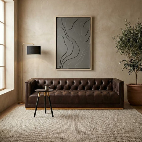 A dark brown leather Chesterfield sofa with a tufted design, featuring three seats and positioned against a neutral-toned wall. A modern abstract artwork hangs above the sofa, complemented by a black floor lamp and a small side table. A potted plant adds a touch of greenery, while a textured area rug covers the floor. Natural light streams in through large windows, enhancing the warm ambiance of the space
