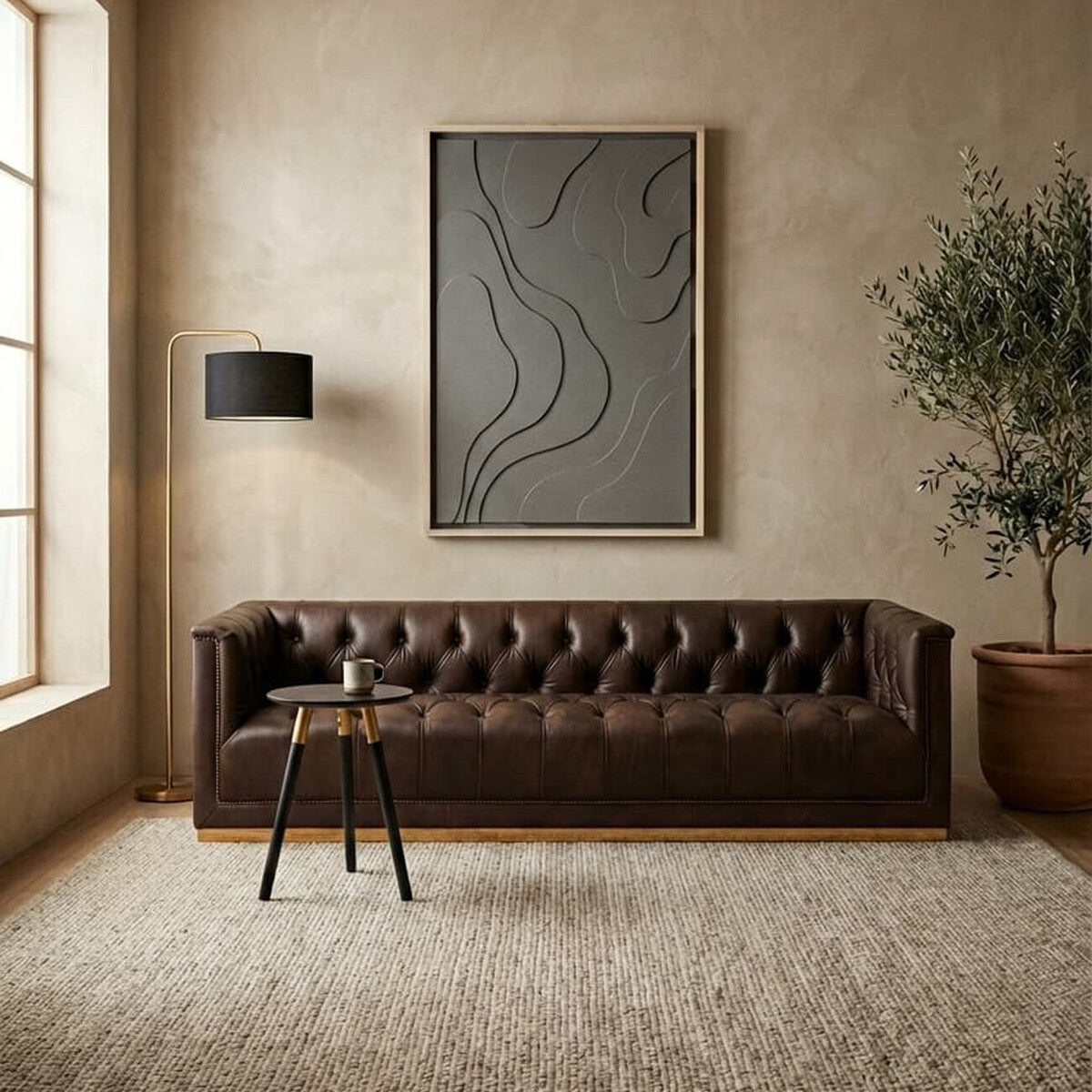 A dark brown leather Chesterfield sofa with a tufted design, featuring three seats and positioned against a neutral-toned wall. A modern abstract artwork hangs above the sofa, complemented by a black floor lamp and a small side table. A potted plant adds a touch of greenery, while a textured area rug covers the floor. Natural light streams in through large windows, enhancing the warm ambiance of the space