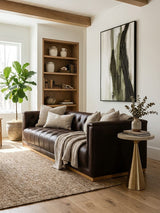 A dark brown leather three-seater Chesterfield sofa is positioned in a cozy living room setting. The sofa features classic tufted upholstery and is adorned with several decorative pillows and a soft throw blanket draped over one arm. A round side table with a plant and a textured rug complement the sofa, while a bookshelf filled with decorative items and a large piece of wall art are visible in the background. Natural