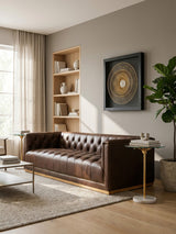 A dark brown leather Chesterfield sofa with a tufted backrest and armrests, positioned in a well-lit living room. The sofa is complemented by a light-colored area rug and a small side table with a decorative lamp. A bookshelf with wooden shelves and a framed artwork are visible in the background, along with a potted plant. Natural light streams in through large windows, creating a warm and inviting