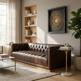 A modern three-seater Chesterfield sofa in dark brown leather, featuring tufted upholstery and a sleek design. The sofa is positioned in a well-lit living room with large windows, a light-colored rug underneath, a small side table with a lamp, and a decorative shelf in the background. A piece of abstract wall art hangs above the sofa, adding a contemporary touch to the space